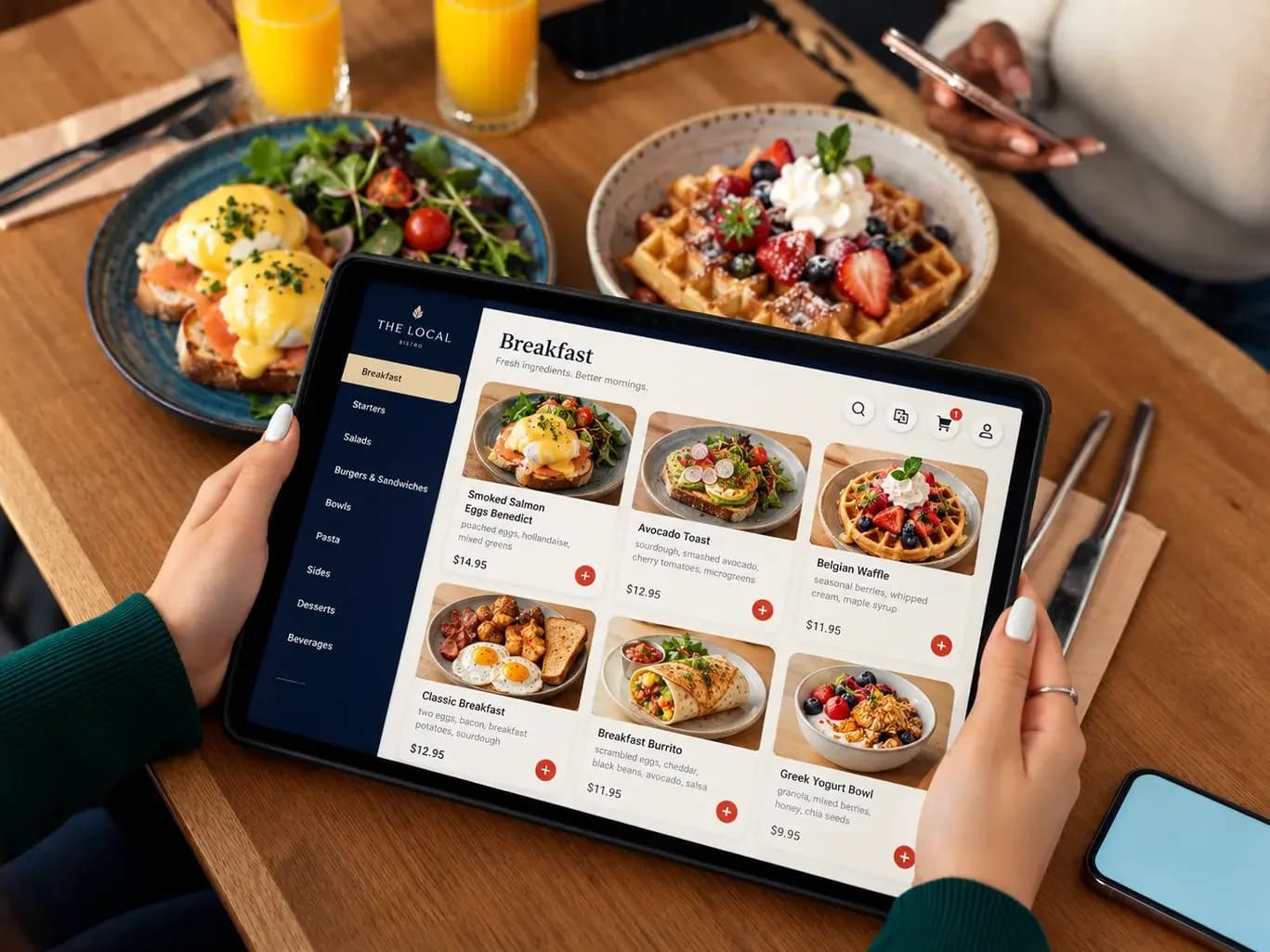 From above, two hands hold a tablet over a breakfast table. The tablet shows a café’s breakfast menu with photos of eggs Benedict, avocado toast and a Belgian waffle, and the same dishes sit on the table around it.