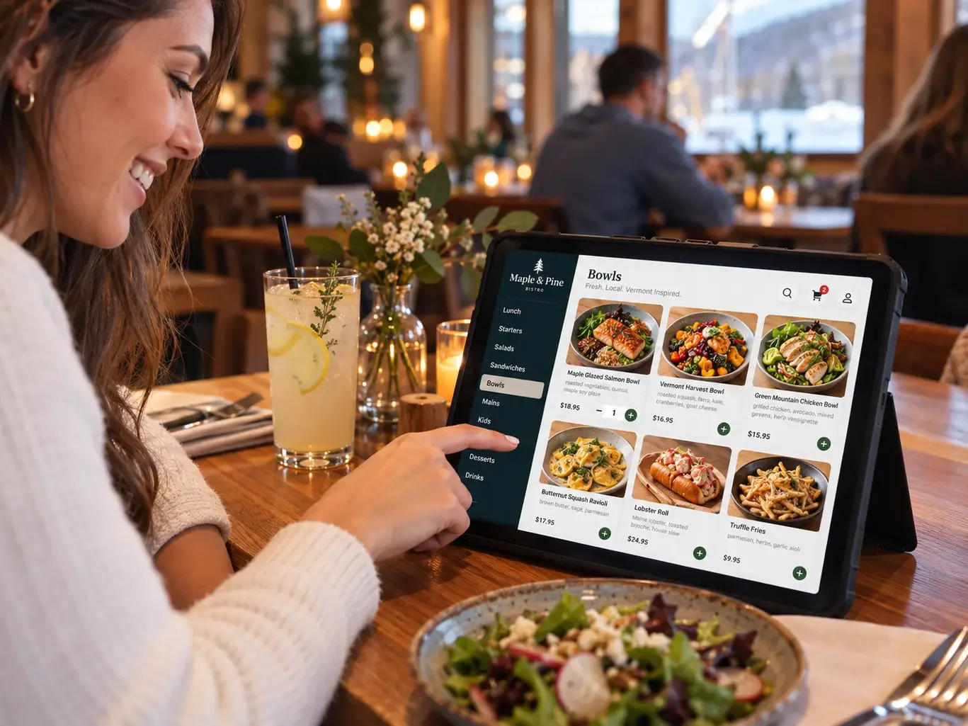 A smiling guest taps a tablet on a stand at a sunny window table. The tablet shows a bistro’s lunch menu of bowls with photos and prices, and one dish has a quantity stepper set to 1.